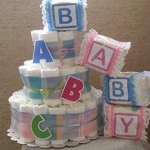 May include: A three-tiered diaper cake constructed from rolled white diapers, wrapped in blue and tan plaid ribbon, and decorated with cutout letters B, A, B, C. Beside it are four additional rolled diapers, each wrapped in lace and labeled with the letters B, A, B, Y.