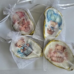 May include: Four small, decorative oyster shells, each featuring a teddy bear design in a hot air balloon or basket. The shells are presented in sheer white drawstring bags with silver ribbon ties. The teddy bears are in shades of pink, blue, and brown.