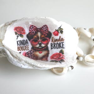 May include: A white oyster shell with a printed graphic of a dog wearing sunglasses and a bow. The text reads "Kinda Boujee Rinda Broke" with red roses. A seashell necklace is next to the shell.