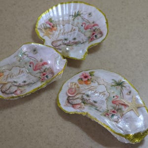 May include: Three decorative oyster shells with gold-painted edges, each featuring a decoupage design of a pink flamingo, palm trees, and the word Florida. One shell includes a small white pearl, while another features a small attached starfish.