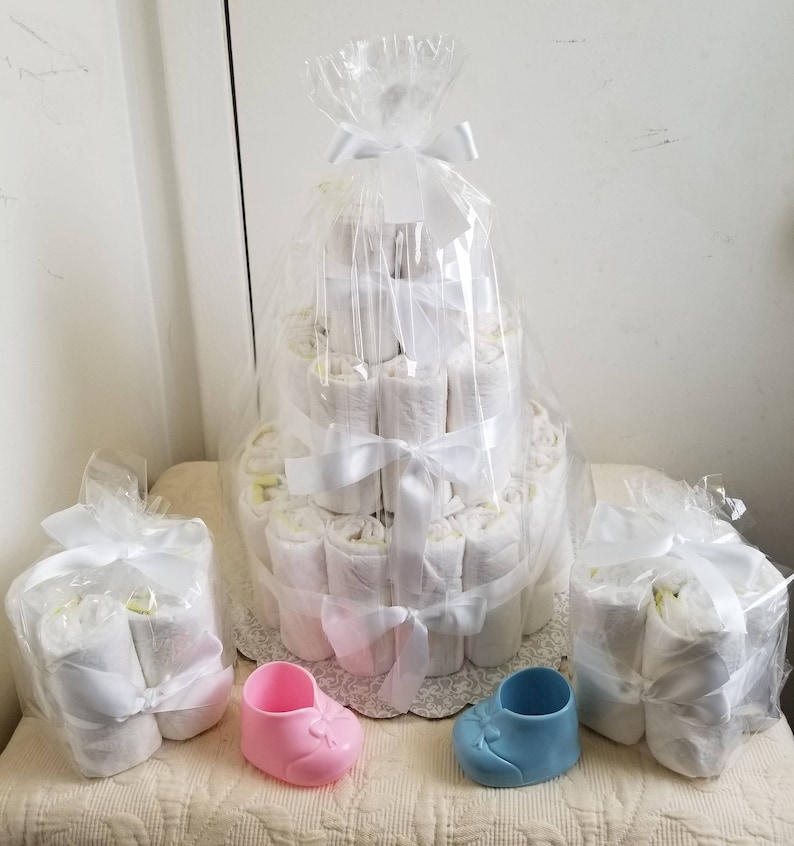 Puede incluir: Un pastel de pa&ntilde;ales de tres niveles envuelto en celof&aacute;n transparente con cintas de sat&eacute;n blanco, acompa&ntilde;ado de dos paquetes m&aacute;s peque&ntilde;os de pa&ntilde;ales enrollados y dos zapatos de beb&eacute; de pl&aacute;stico en miniatura en rosa y azul pastel.