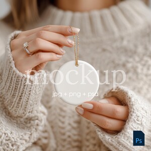 May include: A white, round ornament with a gold chain is held by a person wearing a beige sweater. The ornament has the word "mockup" printed on it, along with the text "jpg + png + psd".