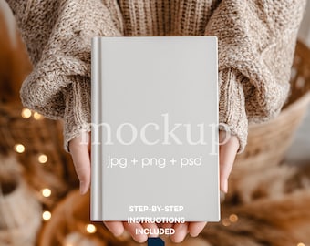 May include: A light gray book mockup held in front of a neutral background. The book has the word "mockup" printed on it, along with "jpg + png + psd". Additional text includes "STEP-BY-STEP INSTRUCTIONS INCLUDED" and "SMART OBJECT".