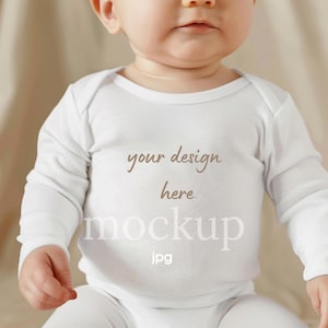 Op de afbeelding: Witte babybody met lange mouwen. De tekst "your design here mockup jpg" is in bruin en wit op de voorkant gedrukt. De baby draagt de body.