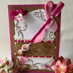 May include: A handmade greeting card with a pink border and brown and white layers. The card is decorated with pink ribbons, flowers, and a butterfly. The layers have decorative floral patterns and lace trim.