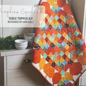 May include: A table topper kit featuring a patchwork quilt with a pumpkin theme. The quilt is made of various fabrics in shades of orange, red, green, yellow, and blue. The quilt is bordered with a solid orange fabric and has a white background. The text "Pumpkins Squared TABLE TOPPER KIT DESIGNED BY LORI HOLT" is printed on a white banner.