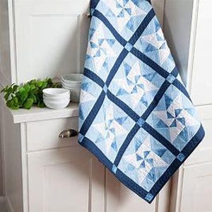 May include: A blue and white quilt with a geometric pattern. The quilt is made of squares and triangles, and the pattern is reminiscent of a pinwheel. The quilt is hanging on a white wall.