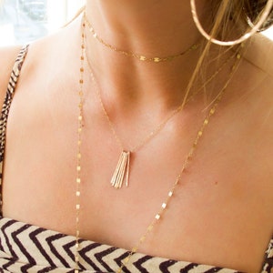 Könnte beinhalten: Eine Frau trägt eine goldene Halskette mit einer zarten Kette und einem Anhänger aus mehreren dünnen Goldstäben. Die Halskette ist über eine zweite goldene Kette mit kleinen, runden Gliedern gelegt. Die Frau trägt ein braunes und weiß gestreiftes Oberteil.
