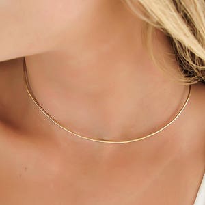 May include: A simple gold choker necklace on a person's neck. The necklace is thin and delicate, and it is made of a smooth metal.