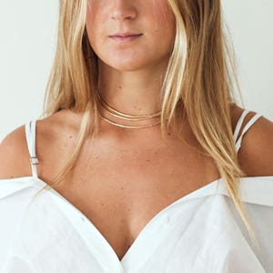 May include: A woman wearing a white off-the-shoulder shirt with a gold layered choker necklace.