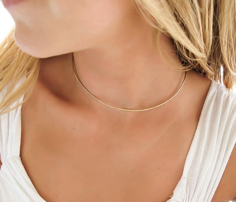 May include: A simple gold choker necklace on a person's neck. The necklace is thin and delicate, and it is made of a smooth metal.