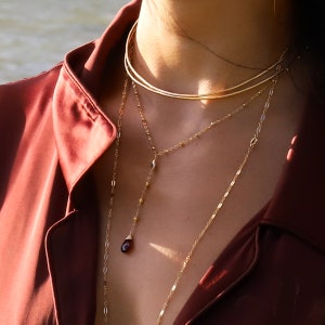 May include: A layered gold necklace with a small, dark red gemstone pendant. The necklace is worn by a person with a red shirt.