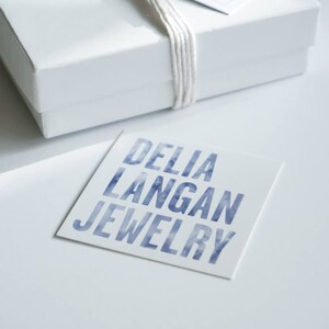 May include: A white box with a white string tied around it. The box is sitting on a white surface. A white card with the text "DELIA LANGAN JEWELRY" in blue lettering is in front of the box.