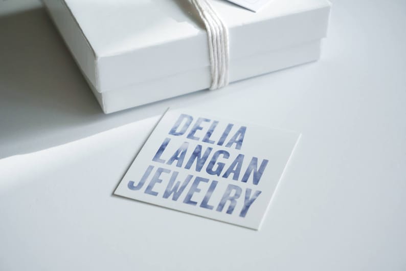 May include: A white box with a white string tied around it. The box is sitting on a white surface. A white card with the text "DELIA LANGAN JEWELRY" in blue lettering is in front of the box.