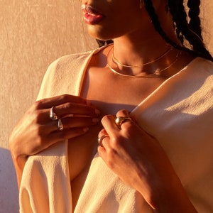 May include: A person wearing a light beige top with a gold chain necklace and multiple gold rings on their fingers.