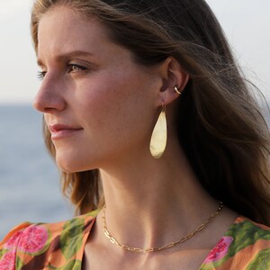 May include: Gold teardrop earrings with a hammered texture. The earrings are worn by a person with long brown hair and a gold chain necklace.