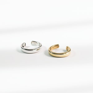 May include: Two ear cuffs, one silver and one gold, are shown on a white surface. The ear cuffs are simple and have a rounded shape.