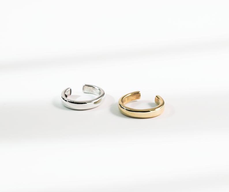 May include: Two ear cuffs, one silver and one gold, are shown on a white surface. The ear cuffs are simple and have a rounded shape.