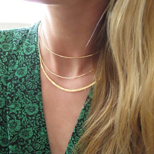 May include: Three gold necklaces layered on a person's neck. The necklaces are different widths and have a shiny finish.