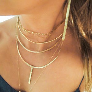 May include: A layered gold necklace set with a hammered pendant. The necklace set features a variety of chain styles, including a delicate chain, a thicker chain, and a hammered chain.