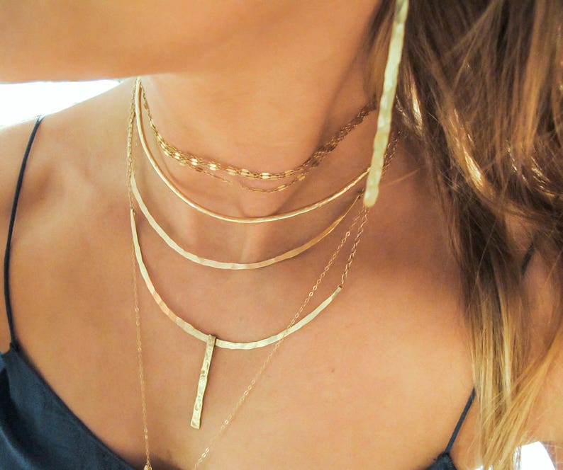 May include: A layered gold necklace set with a hammered pendant. The necklace set features a variety of chain styles, including a delicate chain, a thicker chain, and a hammered chain.