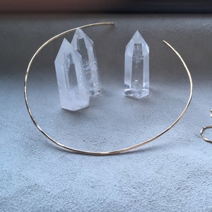 May include: A gold choker necklace with a hammered finish. The necklace is laying on a gray surface with three clear quartz crystals.