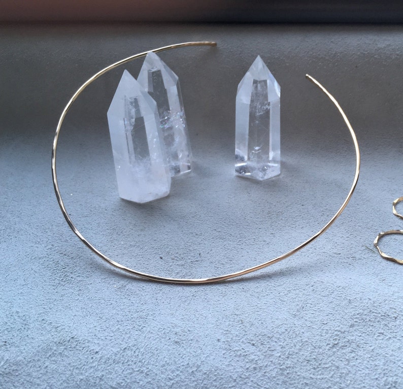 May include: A gold choker necklace with a hammered finish. The necklace is laying on a gray surface with three clear quartz crystals.
