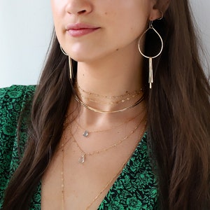 May include: A woman wearing a green floral dress with a gold necklace and earrings. The necklace is layered with several chains and features a small blue gemstone pendant.