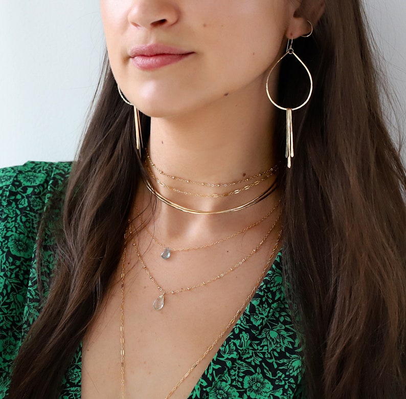 May include: A woman wearing a green floral dress with a gold necklace and earrings. The necklace is layered with several chains and features a small blue gemstone pendant.