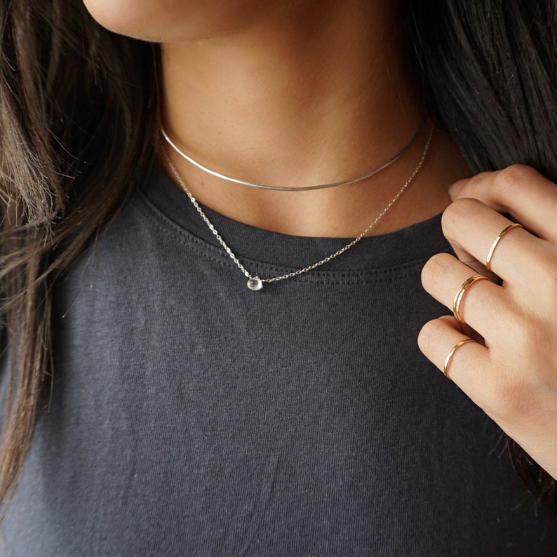 May include: A silver necklace with a small, clear gemstone pendant. The necklace is layered over a silver choker.