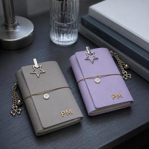 May include: Two rectangular notebooks, one grey and one lavender, each featuring a gold-tone star clip, a thin gold elastic band with a circular charm marked with the letter P, and the gold-tone initials PM embossed on the front cover.