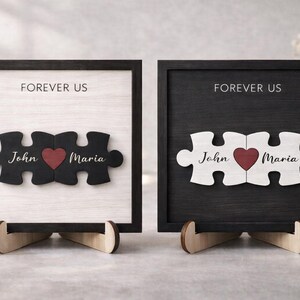 May include: Two framed wooden puzzle pieces, one black and one white, each with a heart and names "John" and "Maria." The text "FOREVER US" is above the puzzle pieces. The frames are displayed on wooden stands.