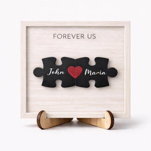 May include: A framed wooden puzzle with the words "Forever Us" above three puzzle pieces. The puzzle pieces are black with the names "John" and "Maria" and a red heart. The frame is white and sits on a wooden stand.