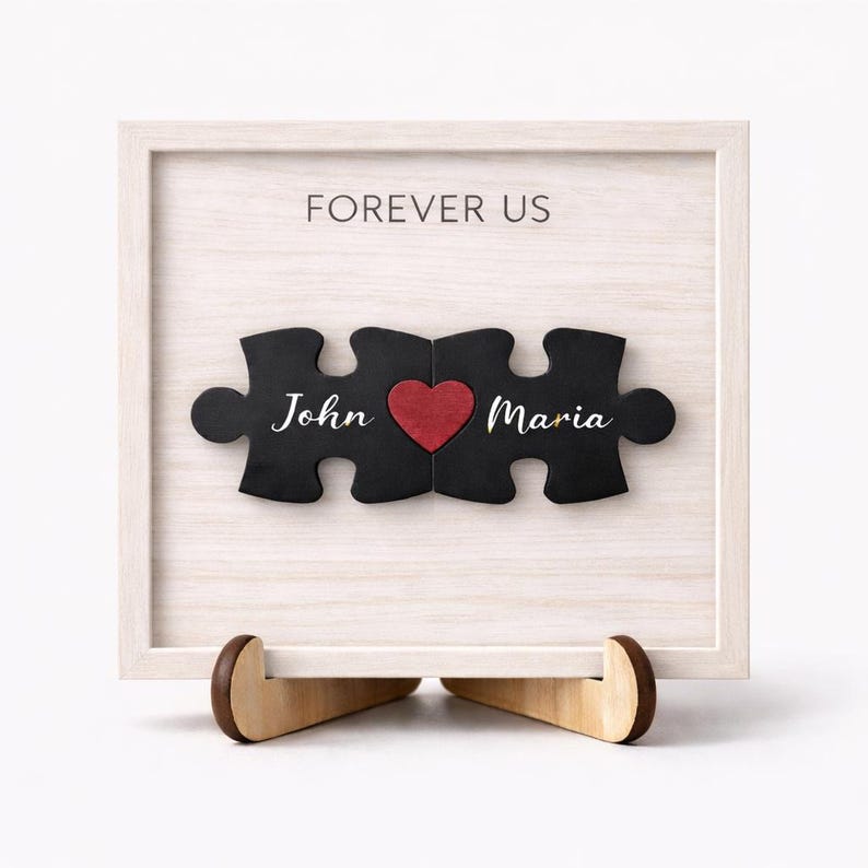 May include: A framed wooden puzzle with the words "Forever Us" above three puzzle pieces. The puzzle pieces are black with the names "John" and "Maria" and a red heart. The frame is white and sits on a wooden stand.