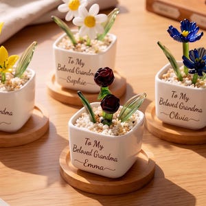 May include: Small white ceramic pots with glass flowers, including daisies, roses, and blue flowers. Each pot has a message, "To My Beloved Grandma" followed by a name. The pots sit on small wooden coasters, creating a charming display.