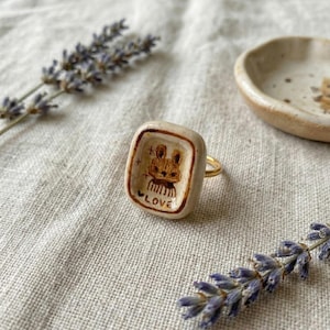 以下が含まれることがあります： 手作りの指輪。四角いオフホワイトのセラミックの表面に、手描きのウサギのイラストと「LOVE」の文字が描かれています。指輪はゴールドカラーのバンドで、ニュートラルなリネンの背景に、ラベンダーの小枝が添えられています。