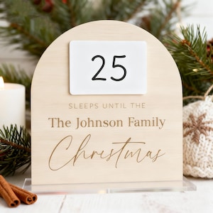 Könnte beinhalten: Ein hölzerner Weihnachtskalender mit einer weißen Karte mit der Zahl 25. Der Text auf dem Kalender lautet "SLEEPS UNTIL THE The Johnson Family Christmas". Der Kalender ist gewölbt und steht auf einer transparenten Basis.