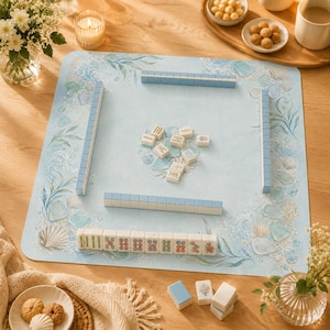 May include: A Mahjong game set with light blue tiles and racks, arranged on a sea-themed mat. The mat features ocean wave and seashell illustrations. The game is set on a wooden table, with snacks and flowers in the background.