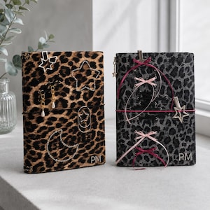Può includere: Due diari rettangolari con copertina leopardata. Il diario a sinistra presenta motivi a stella e luna in strass, ciondoli dorati e le iniziali PM. Il diario a destra ha una stampa leopardata grigia, lacci in nastro rosa e una stella.