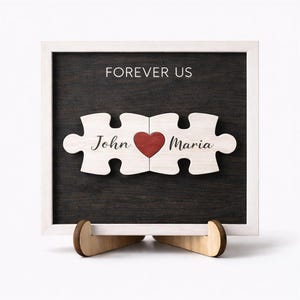 May include: A wooden puzzle piece sign with the words "Forever Us" and the names "John" and "Maria". The puzzle pieces are connected with a red heart in the center. The sign is in a white frame and sits on a wooden stand.