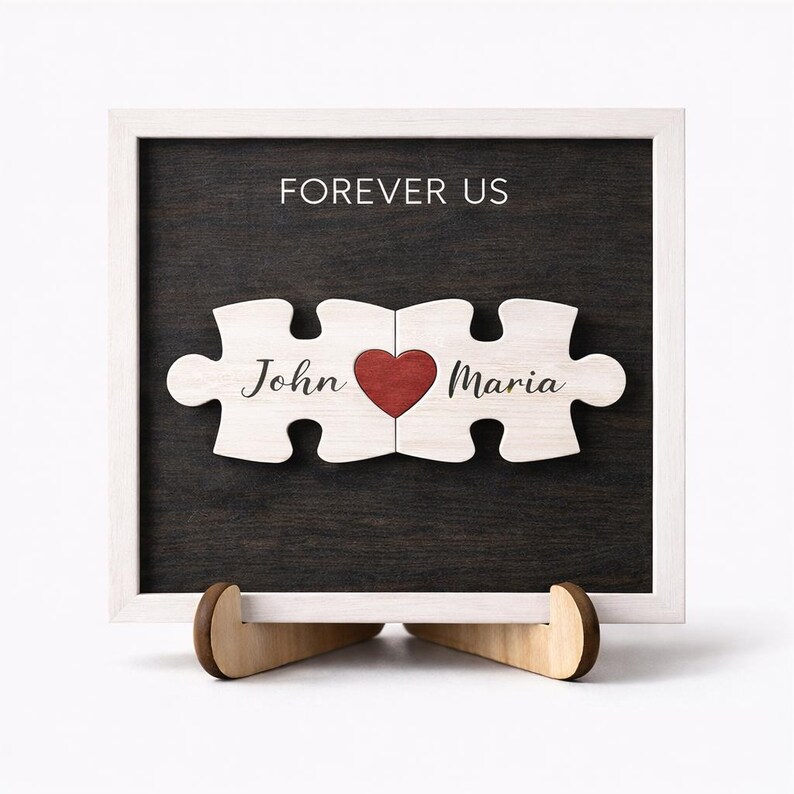 May include: A wooden puzzle piece sign with the words "Forever Us" and the names "John" and "Maria". The puzzle pieces are connected with a red heart in the center. The sign is in a white frame and sits on a wooden stand.