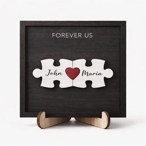 May include: A wooden framed sign with the words "FOREVER US" at the top. Two puzzle pieces with the names "John" and "Maria" and a red heart in the middle. The sign is displayed on a wooden stand.