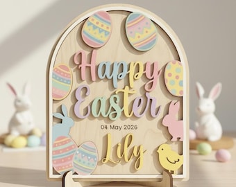 Peut inclure: Panneau de Pâques en bois avec l'inscription "Happy Easter" en couleurs pastel, ainsi que la date "04 mai 2026" et le nom "Lily". Le panneau présente des œufs décoratifs, des lapins et un poussin.