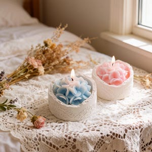 May include: Two lit flower-shaped candles, one blue and one pink, sit on a white lace tablecloth. The candles are in white, textured holders. Dried flowers and a bed are in the background, near a window.