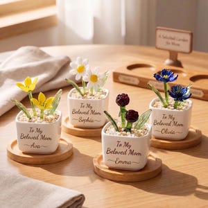 Puede incluir: Cuatro pequeñas macetas de cerámica blanca con flores de cristal, cada una sobre un posavasos de madera. Las macetas tienen flores de diferentes colores: amarillo, blanco, rojo y azul. Cada maceta tiene el texto "To My Beloved Mom" y un nombre.