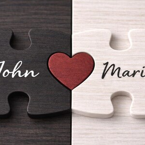 May include: Two wooden puzzle pieces, one black and one white, interlock with a red heart in the center. The black piece says "John" in white script, and the white piece says "Maria" in black script. A romantic gift.