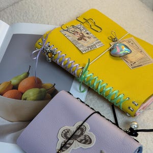 May include: A yellow journal with lavender and mint green ribbon lacing, adorned with a silver bear charm and a heart-shaped pendant. A lavender journal features a butterfly sticker, a star-shaped charm, and a small oval pendant on a black cord.