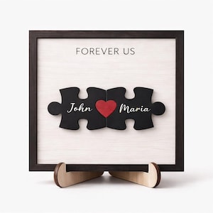 May include: A framed wooden artwork featuring two black puzzle pieces with the names "John" and "Maria" and a red heart. The words "FOREVER US" are printed above. The frame is dark brown, and the artwork is displayed on a wooden stand.