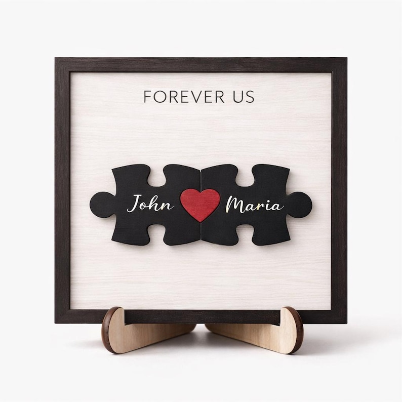 May include: A framed wooden artwork featuring two black puzzle pieces with the names "John" and "Maria" and a red heart. The words "FOREVER US" are printed above. The frame is dark brown, and the artwork is displayed on a wooden stand.