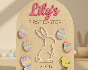 Peut inclure: Panneau en bois sur le thème de Pâques avec le nom "Lily's" en script rose. Le panneau présente une silhouette de lapin, des œufs de Pâques colorés, un poussin jaune et le texte "FIRST EASTER" et "2026". Le panneau est soutenu par un support en bois.